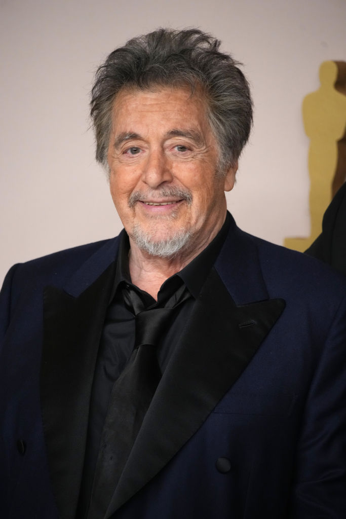 Al Pacino at the 96th Annual Academy Awards smiling while wearing a black shirt and tie and a navy blue suit jacket