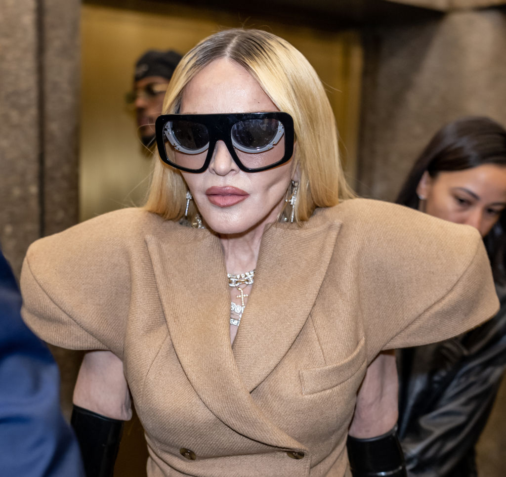 Madonna looks serious as she exits a building wearing large dark sunglasses and a tan suit with oversized shoulders