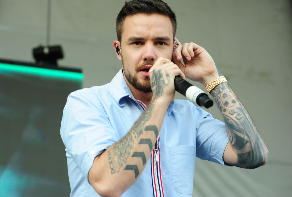 Liam Payne performing on stage in a blue shirt, holding a microphone and touching his earpiece