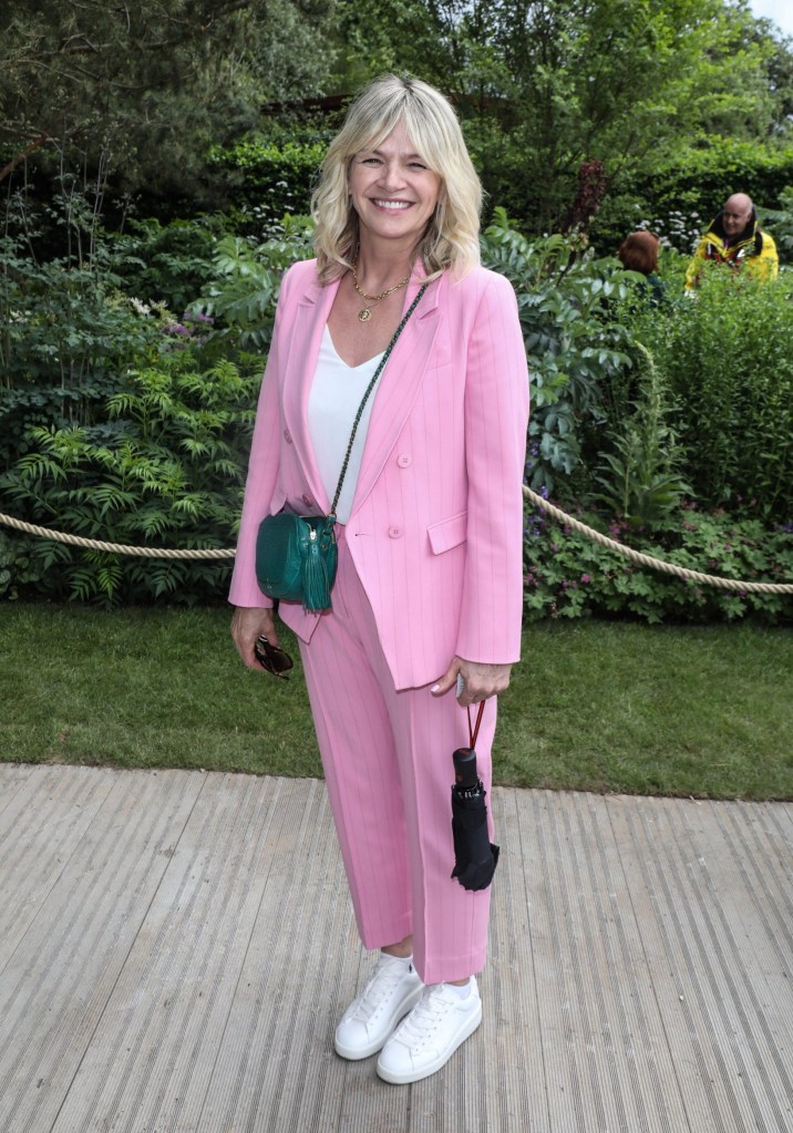 Zoe Ball in a pink suit and white shoes on a deck with greenery in the background