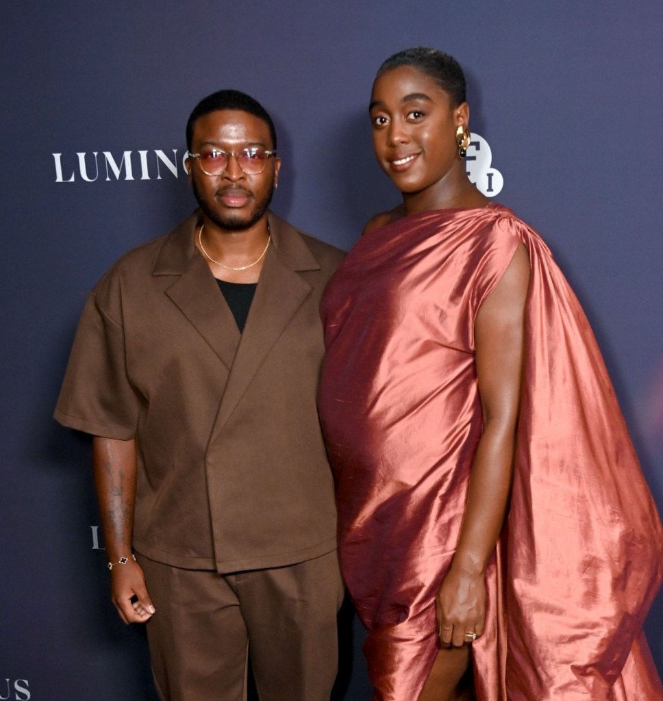 Zackary Momoh and Lashana Lynch on the red carpet in formal clothes, a brown suit and copper gown