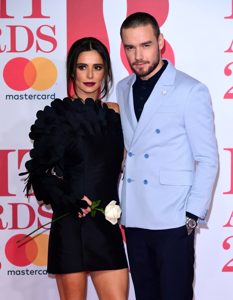 Liam Payne and Cheryl posing together at a red carpet event. Cheryl is wearing a black statement dress while Liam is wearing black trousers, a black shirt, and a pale blue suit jacket