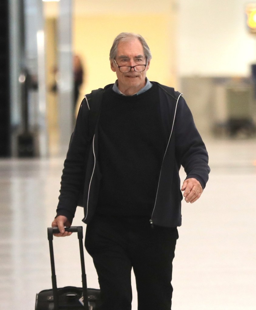 Timothy Dalton walking with a suitcase in an airport wearing glasseson his nose. He is dressed in a dark jacket, top and trousers 