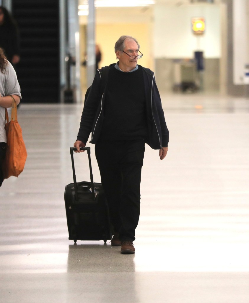 Timothy Dalton walking with a suitcase in an airport wearing glasses on his nose. He is dressed in a dark jacket, top and trousers