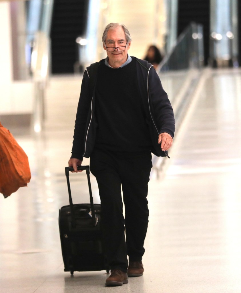 Timothy Dalton walking with a suitcase in an airport wearing glasses on his nose. He is dressed in a dark jacket, top and trousers 