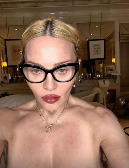 Madonna poses topless for a selfie wearing black-rimmed glasses and a cross necklace,