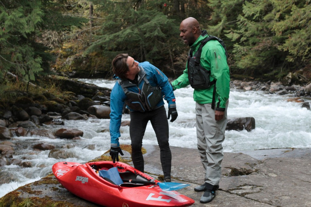 Martin Henderson and Colin Lawrence in a scene from Virgin River, stood by a stream of water with a river rafting boat