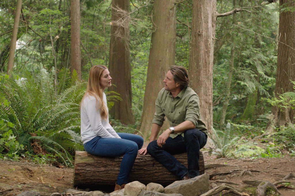 Alexandra Breckenridge and Martin Henderson as Mel and Jack in the woods in Virgin River, sitting on a log in the woods